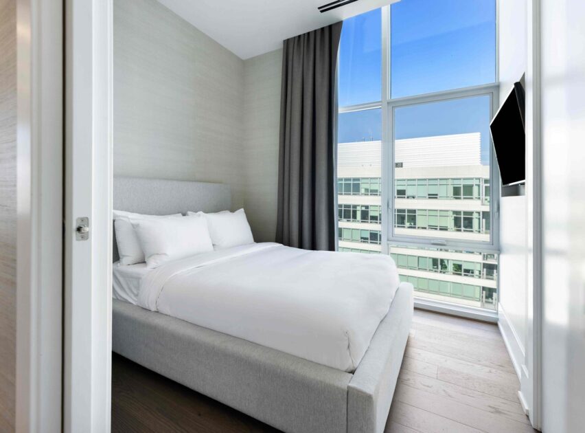 A modern bedroom with a grey upholstered bed, white bedding, large floor-to-ceiling windows, grey curtains, and a wall-mounted TV, overlooking adjacent office buildings under a clear blue sky.