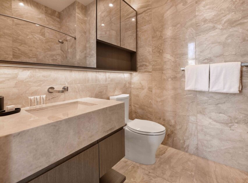 Modern bathroom with beige marble walls and countertop, a white toilet, large mirror cabinets, towel rack with two white towels, and built-in lighting under the cabinets. Minimalist and clean design.
