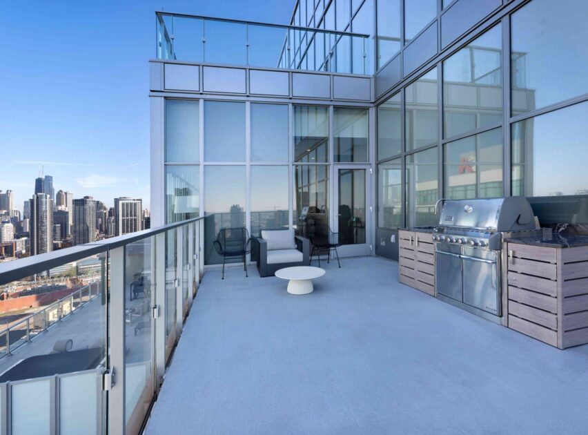 A modern high-rise balcony with glass railings, outdoor seating, a white round table, and a stainless steel grill. The city skyline is visible in the background under a clear blue sky.