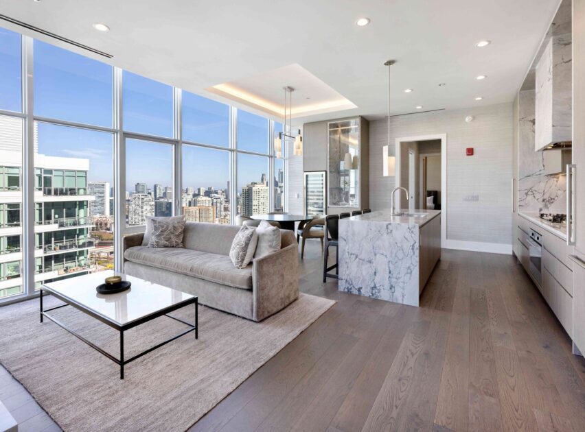 Modern open-concept living room and kitchen with floor-to-ceiling windows, city views, light wood floors, marble countertops, a beige sofa, glass coffee table, and built-in appliances. Bright and airy atmosphere.