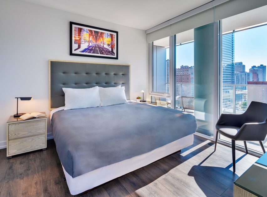 Modern bedroom with a large bed, gray headboard, two nightstands, a chair, and floor-to-ceiling windows offering a city view. A framed cityscape artwork hangs above the bed.