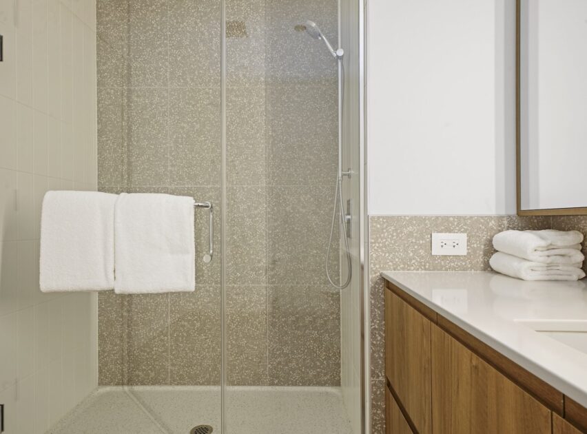 A modern bathroom with a glass shower enclosure, a white towel hanging on the shower door, folded towels on a wooden vanity, and a wall outlet above the counter. The shower features a handheld showerhead.