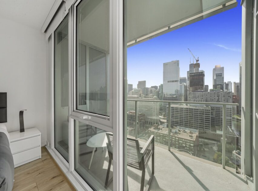 Modern high-rise apartment with floor-to-ceiling windows and a balcony overlooking a city skyline with tall buildings, construction cranes, and clear blue sky. A small table and chair are on the balcony.