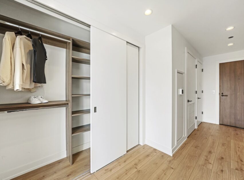 A modern, empty closet with sliding white doors, open to reveal shelves and hanging clothes, in a room with light wood floors, white walls, and a closed wooden entry door.