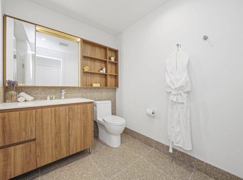 Modern bathroom with a wooden vanity, open shelving, a large mirror, a white toilet, a towel hook with a white bathrobe, and neutral tile flooring and walls. Minimalist and clean design.