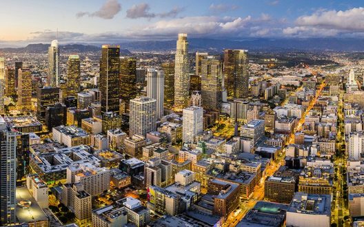Aerial view of a city skyline at dusk, featuring tall skyscrapers with lights on, busy streets, and a mix of modern and older buildings under a partly cloudy sky.