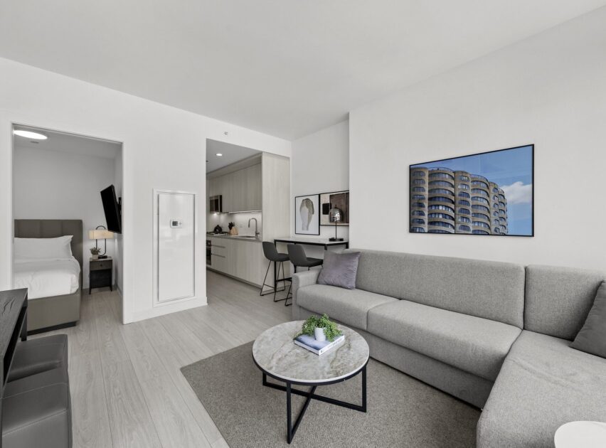 Modern, light-filled apartment living room with a gray sectional sofa, round coffee table, and wall art. Open kitchen and bar seating are visible, along with a glimpse of a bedroom through an open door.