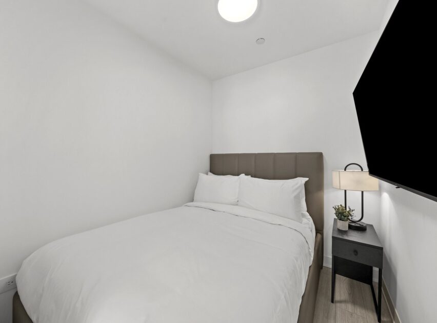 A small, modern bedroom with a double bed, white bedding, a padded headboard, a bedside table with a lamp and small plant, and a wall-mounted TV. The walls are white and the room is simply decorated.