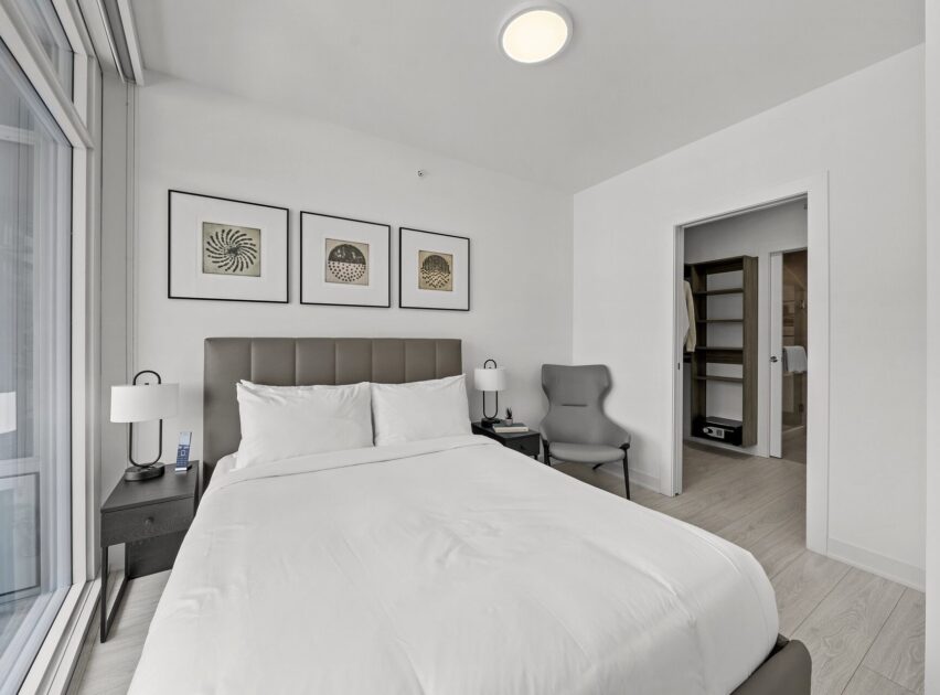 A modern bedroom with a neatly made bed, gray padded headboard, black nightstand, gray armchair, and three framed abstract art pieces on a white wall; an open door reveals a walk-in closet.