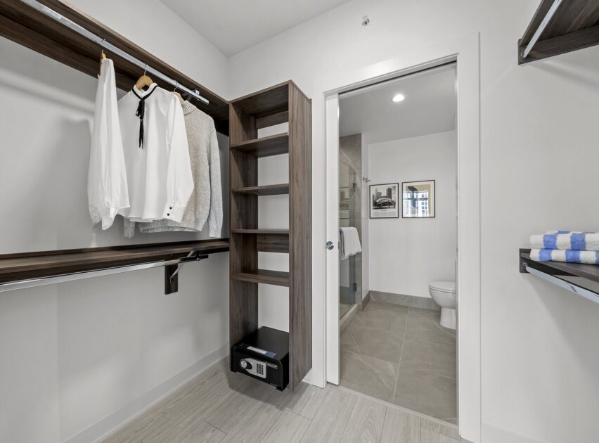 Modern walk-in closet with hanging clothes, open shelves, a small safe, and a doorway leading to a bathroom with a toilet, shower, and framed pictures on the wall.