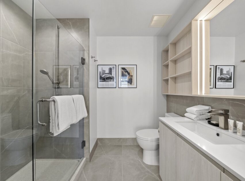 Modern bathroom with glass shower, grey tile flooring and walls, wall-mounted sink with cabinets, large mirror, open shelves with towels, toilet, and framed black-and-white photos on the wall.