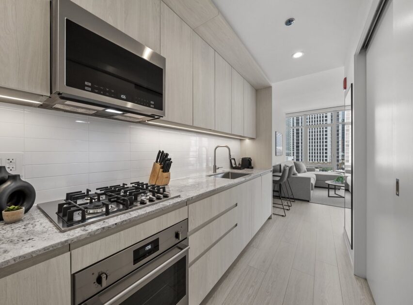 Modern kitchen with light wood cabinets, built-in oven, gas stovetop with knives nearby, white tile backsplash, and a sink. The space opens into a bright living area with large windows and city views.