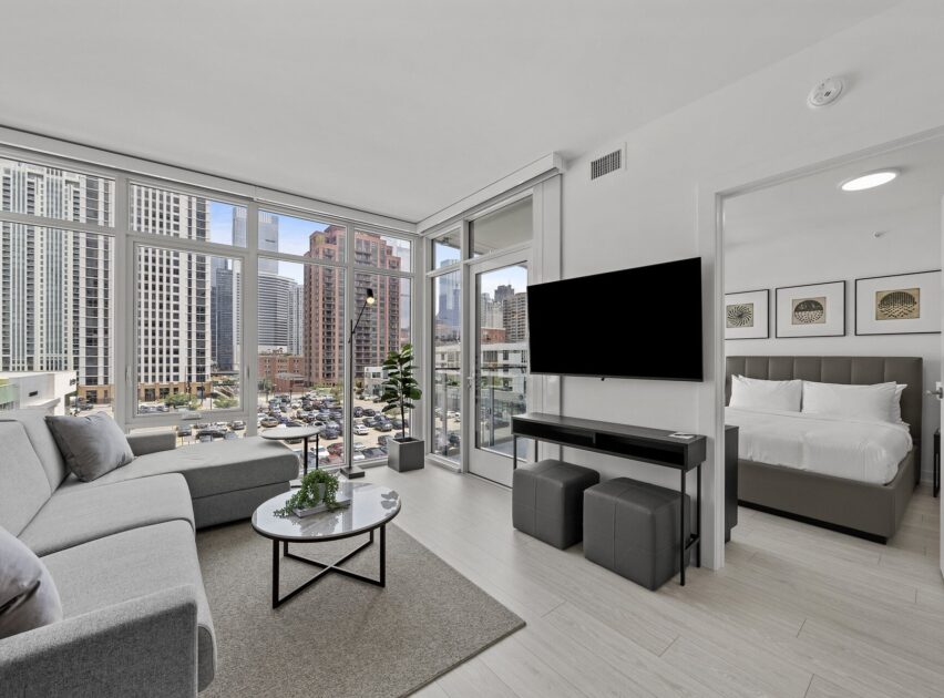 Modern apartment living room with floor-to-ceiling windows, gray sectional sofa, round coffee table, TV on the wall, and a view of city buildings; adjacent bedroom with a neatly made bed is visible.