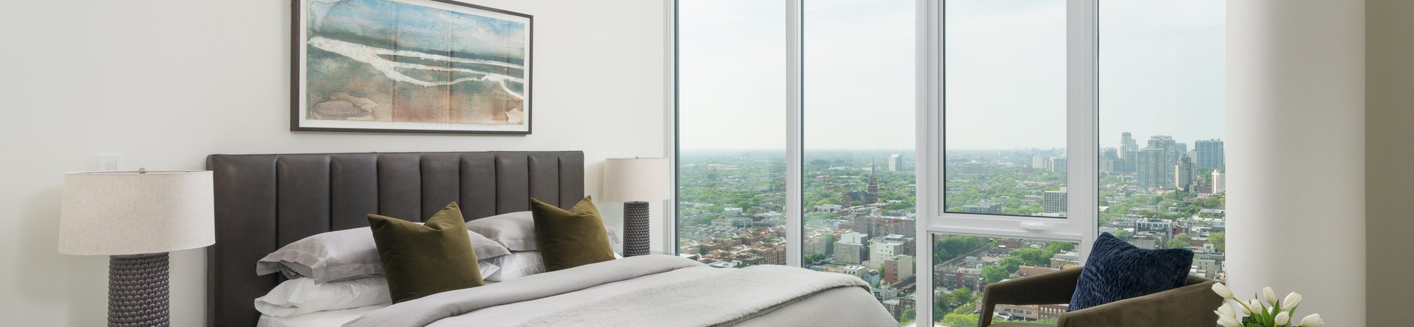 Modern bedroom with large windows offering a cityscape view, a bed with gray and white bedding, two lamps on bedside tables, a piece of abstract art above the headboard, and a vase of flowers on the right.