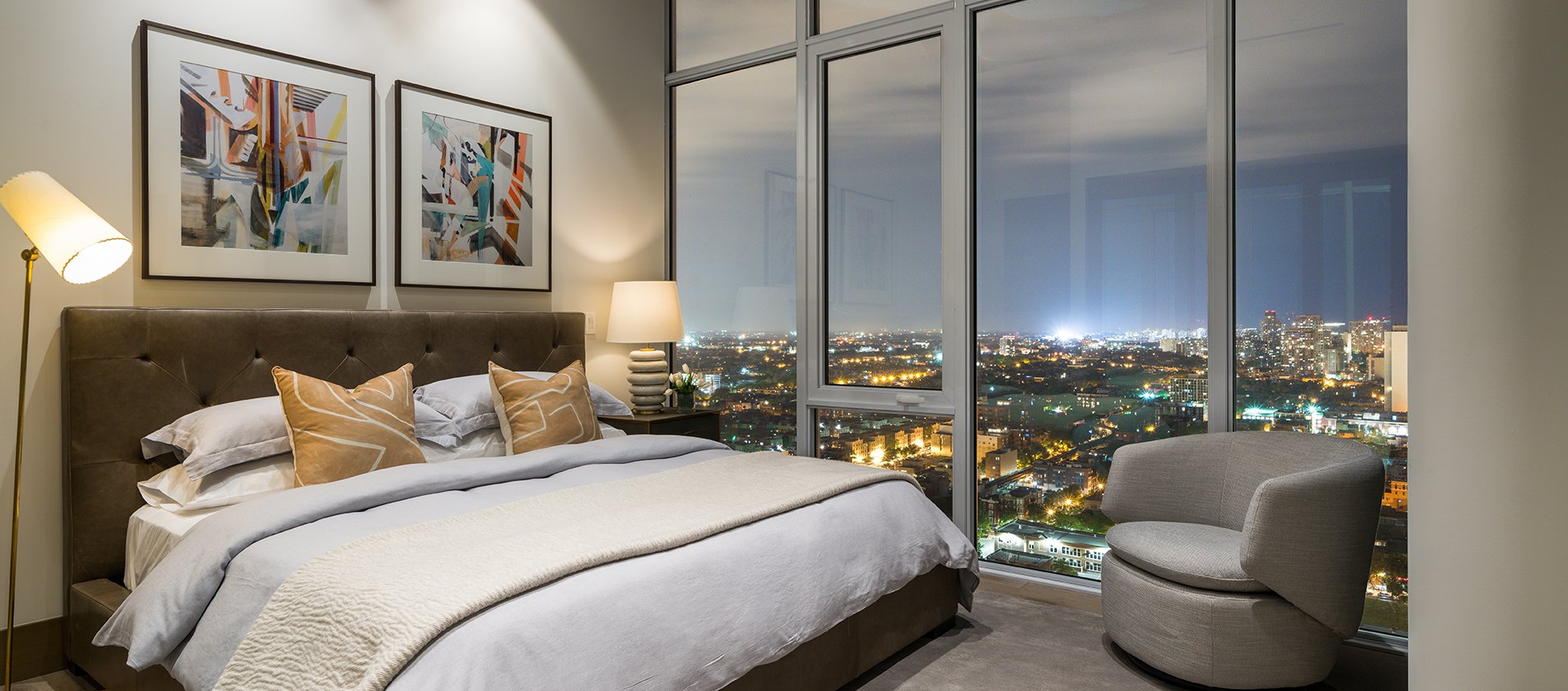 Modern bedroom with a large bed, beige and gray decor, two abstract paintings above the headboard, a floor lamp, armchair, and floor-to-ceiling windows with a nighttime cityscape view.