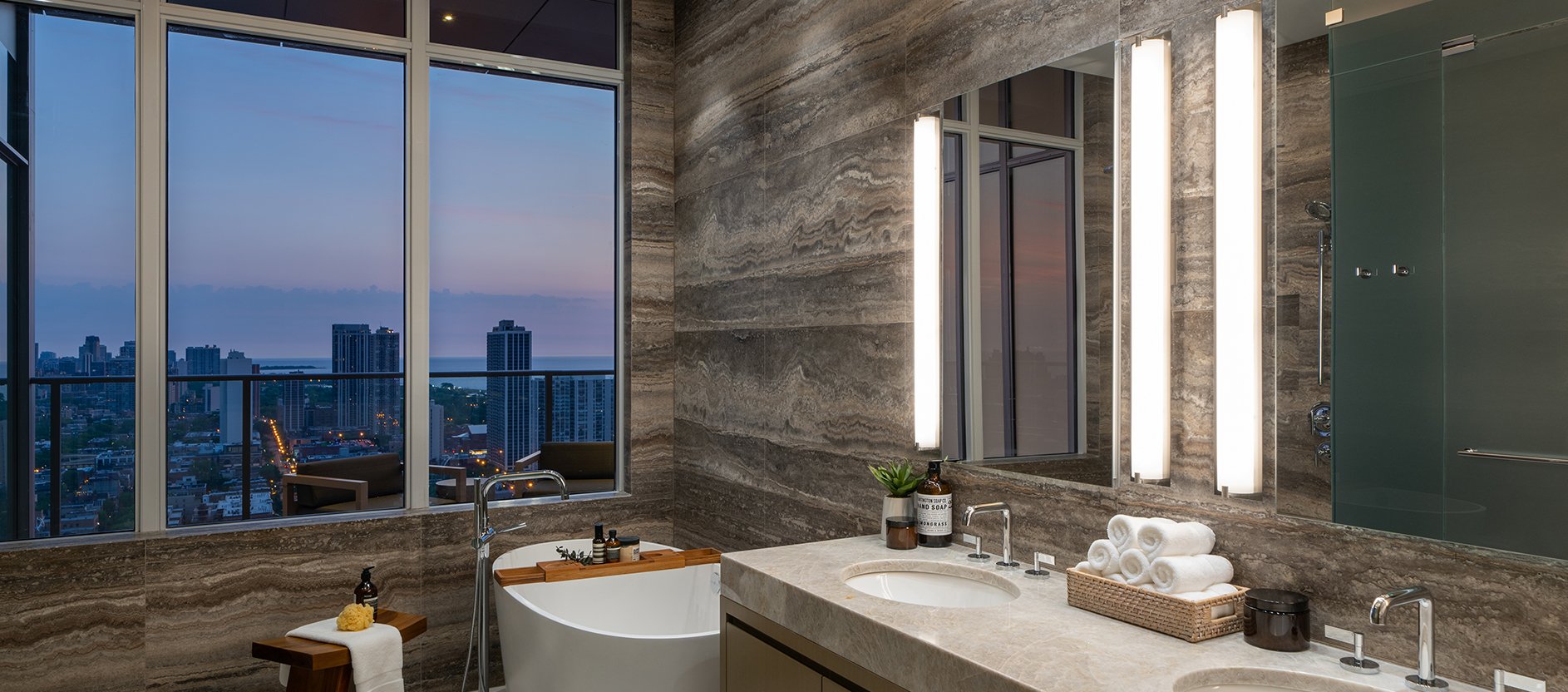 Luxurious bathroom with marble walls, double sinks, a freestanding bathtub, rolled towels, and large illuminated mirrors. Floor-to-ceiling windows provide a city skyline view at sunset.