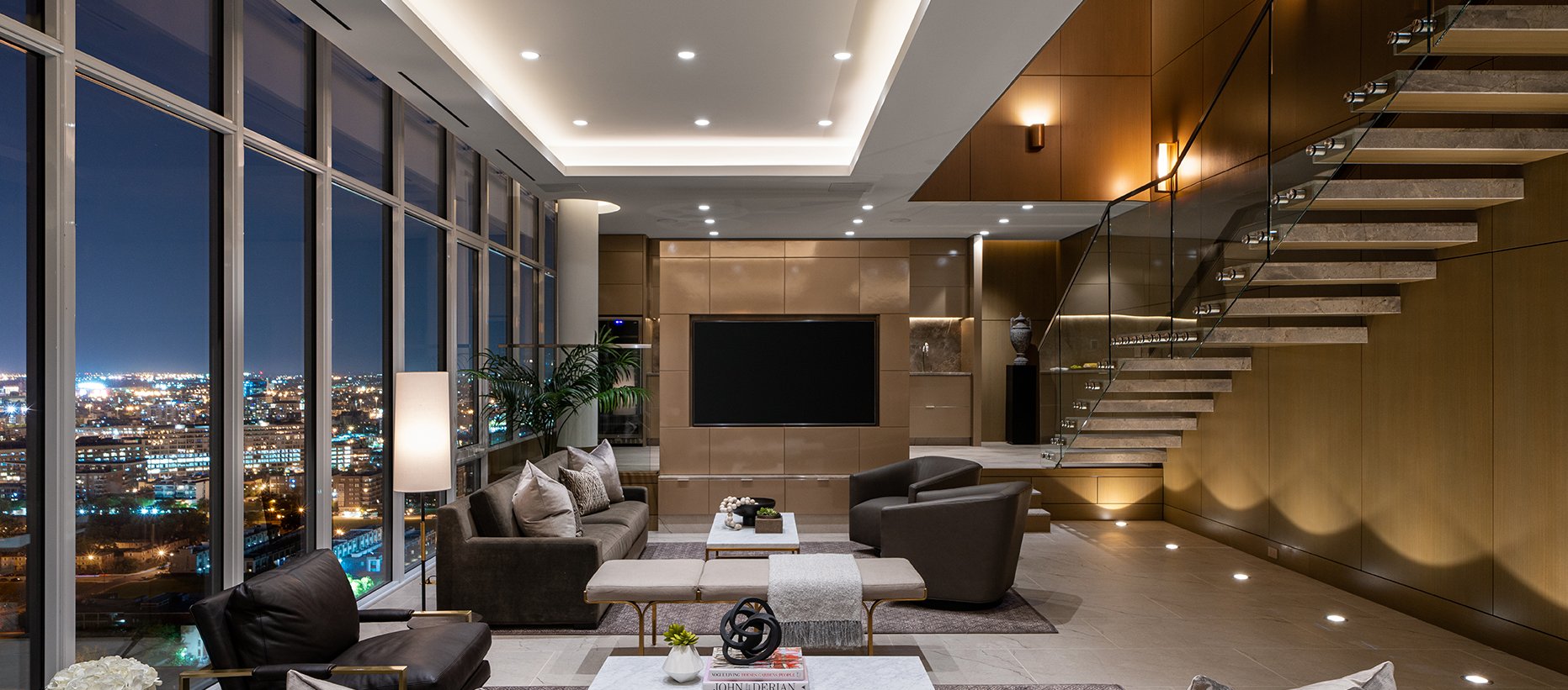 Modern living room with large windows showcasing a city night view, sleek furniture, neutral tones, a wall-mounted TV, plants, and a floating staircase with glass railing leading to an upper level.