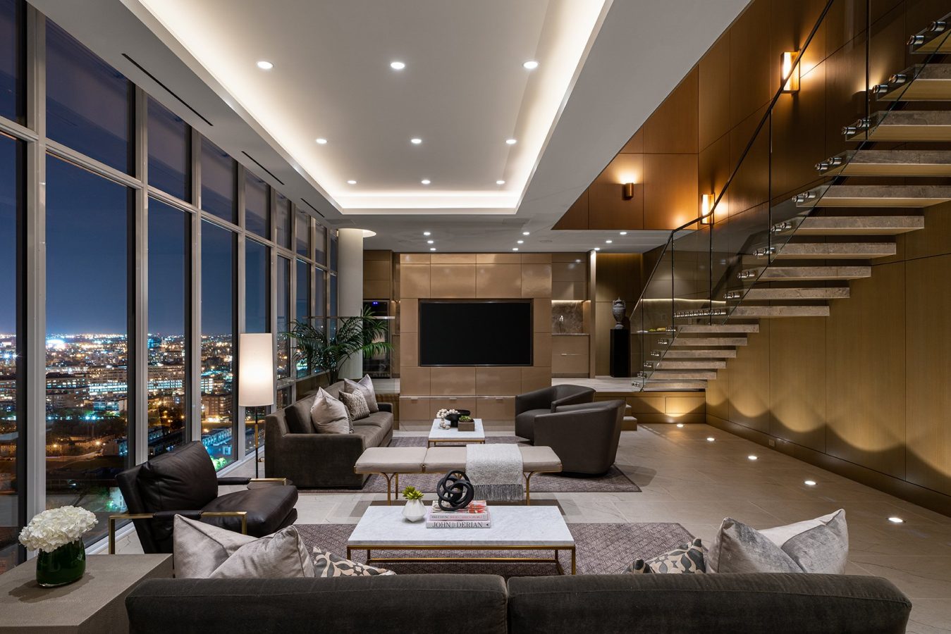 Modern, luxurious living room with floor-to-ceiling windows overlooking a city at night, featuring dark sofas, marble tables, decorative plants, and a floating staircase with glass railings alongside wood-paneled walls.
