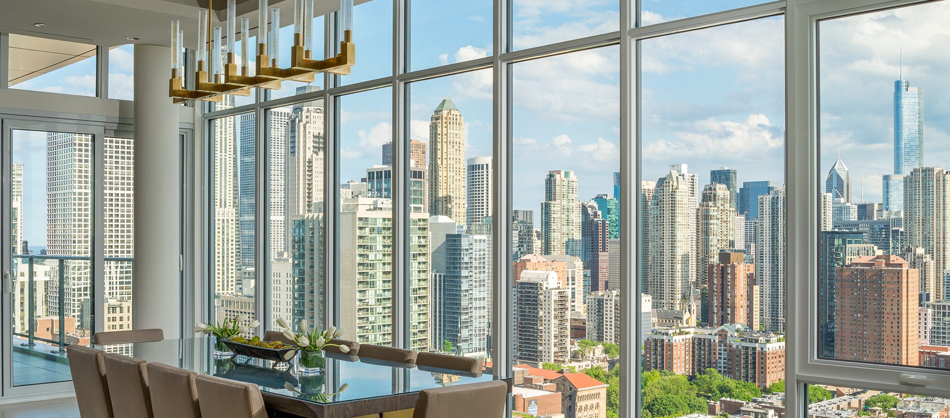 Modern dining area with a large table and elegant chandelier, featuring floor-to-ceiling windows offering a panoramic view of a city skyline with numerous skyscrapers under a partly cloudy sky.