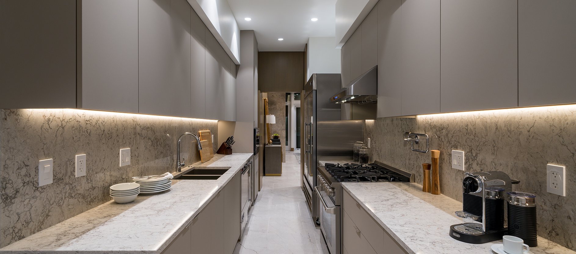 Modern galley kitchen with grey cabinets, white marble countertops and backsplash, under-cabinet lighting, stainless steel appliances, and neatly arranged kitchenware on the counters.