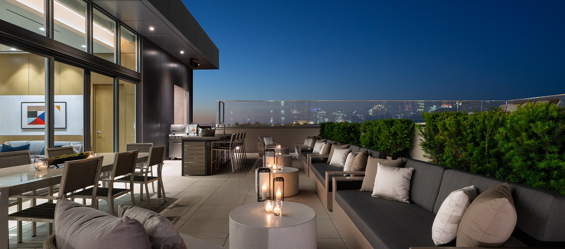 Modern rooftop terrace at dusk with cushioned seating, dining table, potted plants, and city skyline in the background. Lit candles and soft lighting create a cozy, inviting atmosphere. Glass railing offers an unobstructed view.