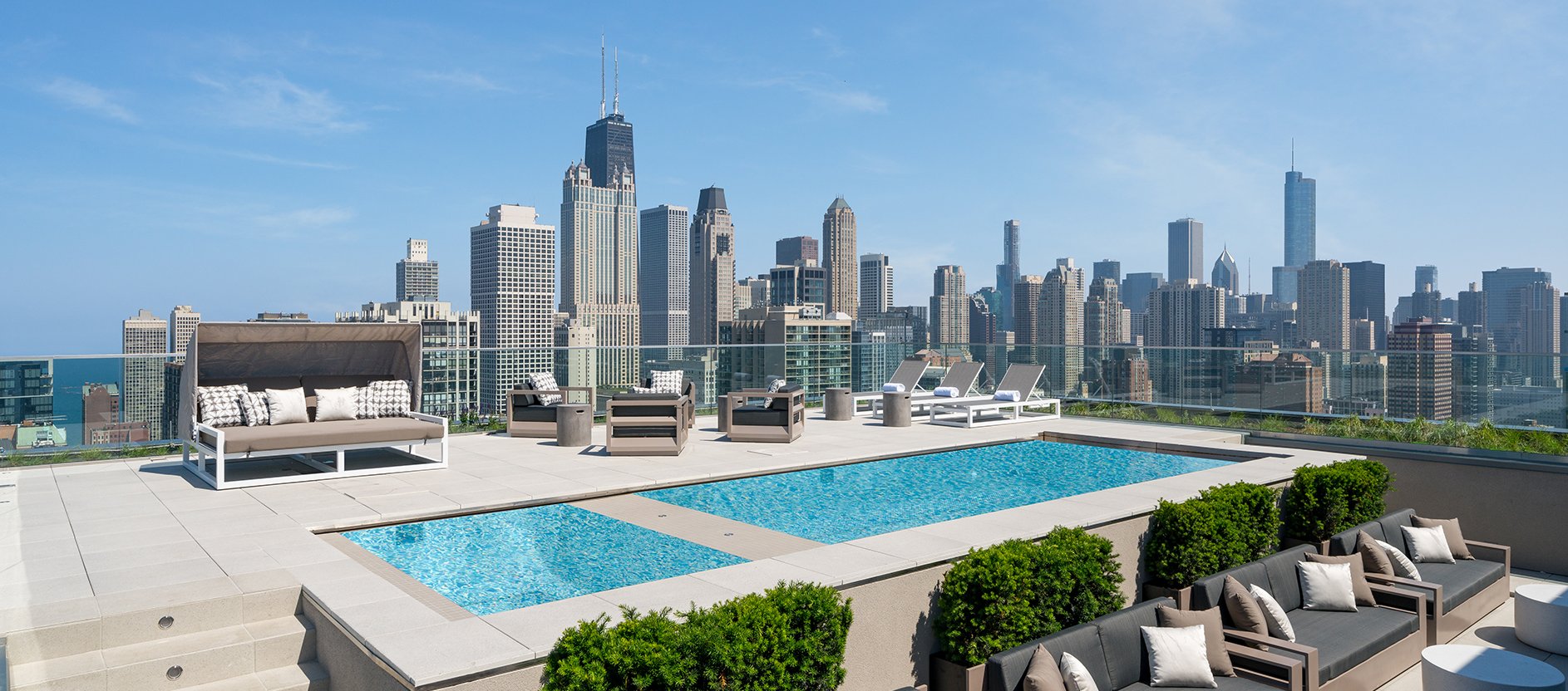 A rooftop pool area with loungers, seating, and greenery overlooks a city skyline with tall skyscrapers under a clear blue sky.