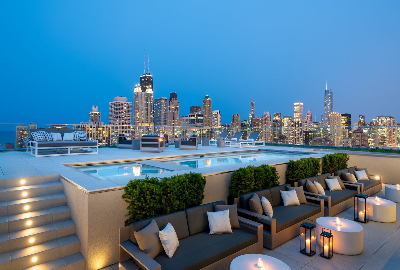 A rooftop lounge with modern seating, lit candles, and a pool overlooks a city skyline at dusk, featuring tall buildings and bright city lights against a clear blue sky.