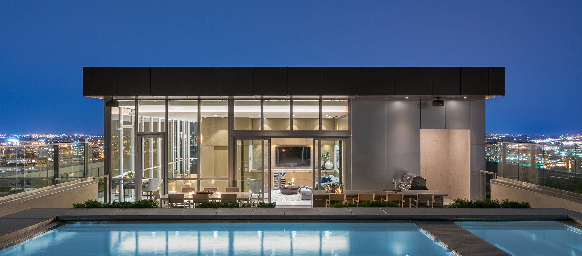 Rooftop lounge with floor-to-ceiling windows, modern outdoor seating, and a swimming pool in the foreground, overlooking a brightly lit cityscape at night.