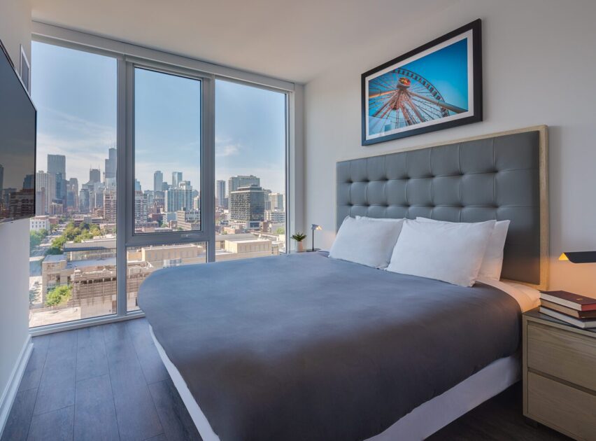 Modern bedroom with a large bed, gray headboard, and gray bedding. Floor-to-ceiling windows reveal a city skyline view. A framed photo hangs above the bed, and a nightstand with books and a lamp is next to it.