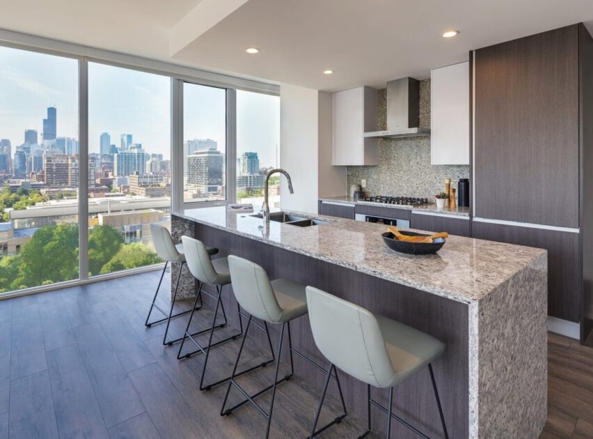 Modern kitchen with a large marble island, four bar stools, built-in appliances, sleek cabinets, and floor-to-ceiling windows offering a view of a city skyline and greenery.
