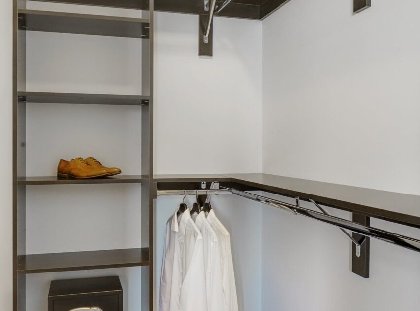 A modern walk-in closet with dark shelves, a pair of brown shoes on one shelf, and several white shirts hanging on hangers from a horizontal rod. The closet has a clean, minimalist look.