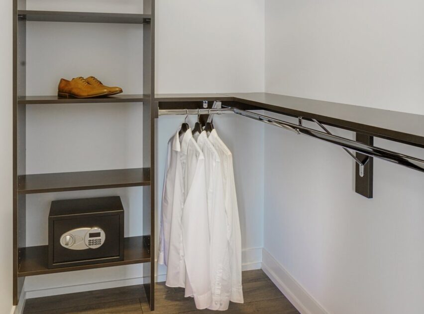 Walk-in closet with dark shelves, a pair of brown dress shoes, a digital safe, and four white shirts hanging neatly on a rail against white walls and a dark floor.