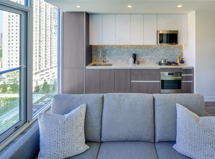 Modern apartment kitchen with light wood and white cabinets, built-in appliances, and a backsplash. In the foreground, a gray sofa with patterned pillows faces the kitchen, and large windows provide a city view.