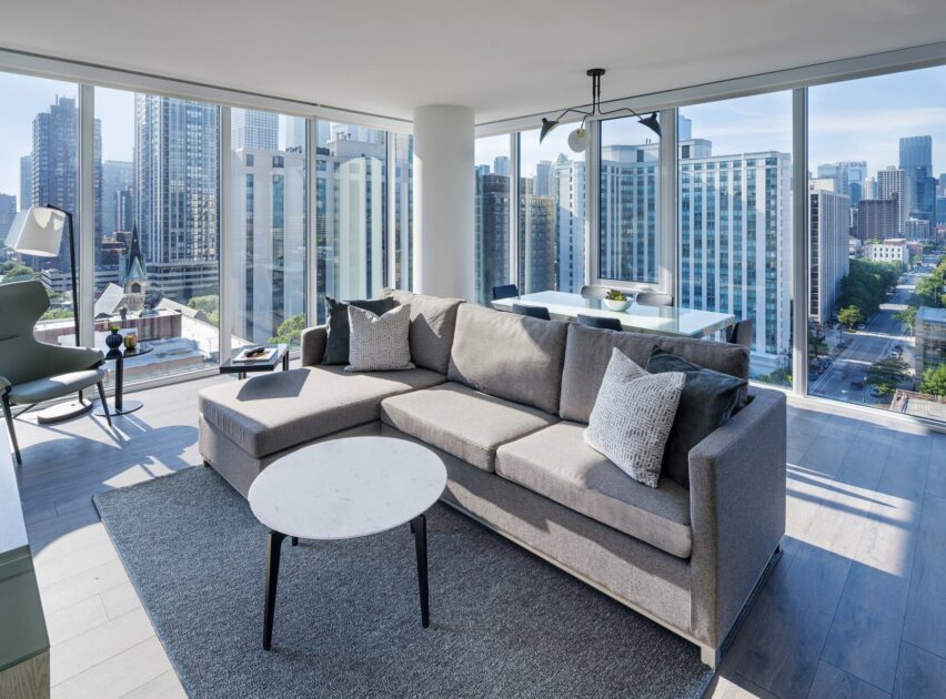 A modern living room with a large gray sectional sofa, marble coffee table, and floor-to-ceiling windows offering a city skyline view. A dining area and contemporary decor are visible in the bright space.