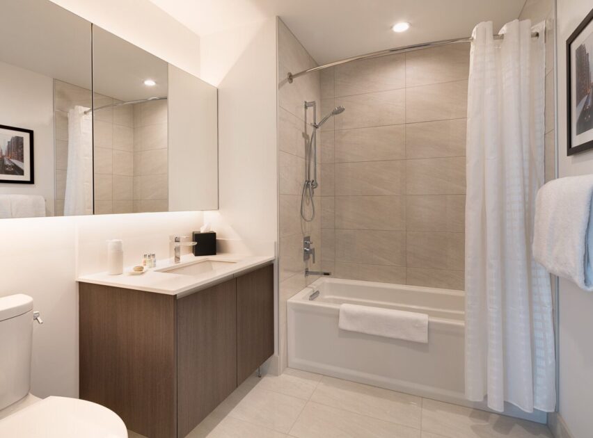 Modern bathroom with light beige tiles, a bathtub with a shower and white curtain, a large mirror above a sink with wooden cabinetry, a toilet, and a towel hanging on the wall. Framed artwork decorates the space.