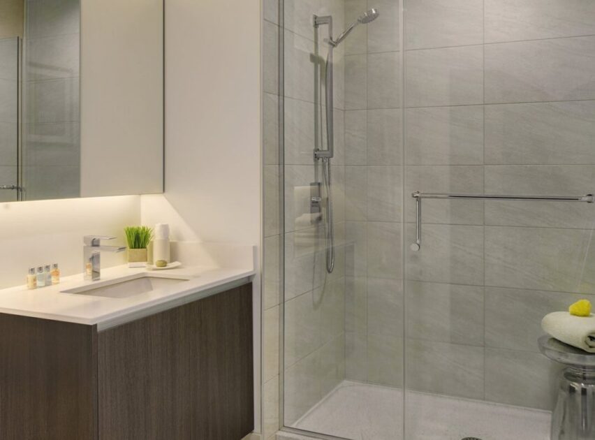 Modern bathroom with a glass-enclosed shower, light gray tiled walls, and a vanity with a white countertop and wood cabinet. The countertop has toiletries, a soap dispenser, and a small plant.