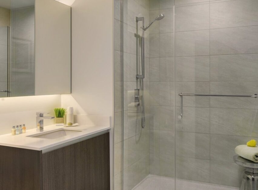 Modern bathroom with a sink and faucet on a wooden vanity, toiletries and green plant on the counter, and a glass-enclosed shower with a wall-mounted showerhead and tiled walls.