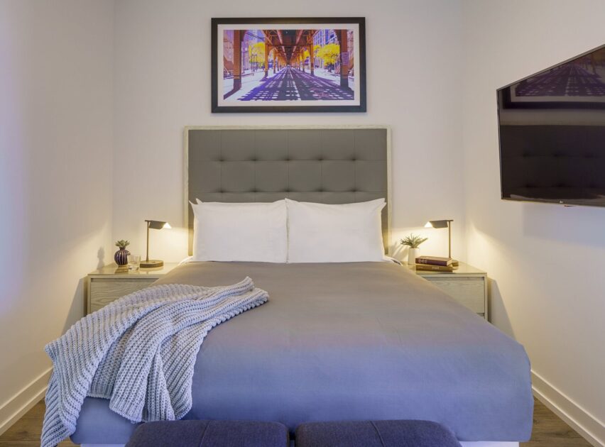 A neatly made bed with gray bedding and a textured blanket sits between two nightstands with lamps. A framed cityscape artwork hangs above the headboard, and a wall-mounted TV is visible on the right.