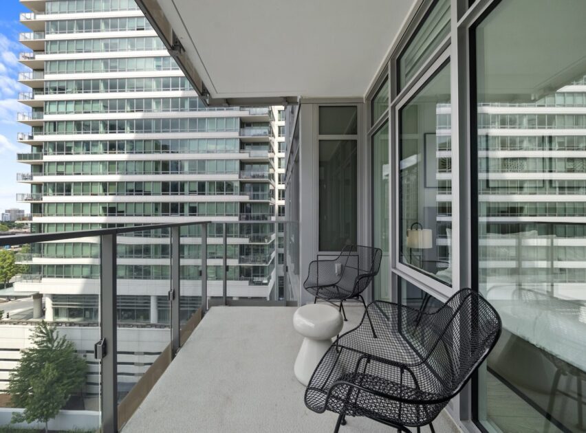 A modern apartment balcony with two black wire chairs, a small white table, and glass railings, overlooking neighboring high-rise buildings with reflective windows.