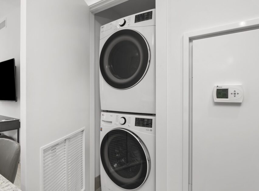 A stacked washer and dryer set is built into a white laundry closet. A thermostat and vent are mounted on the adjacent wall. The space features clean, modern decor with light-colored walls.