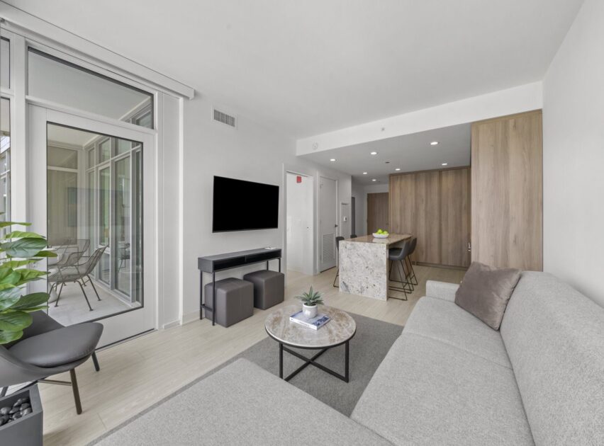 Modern living room with a gray sofa, round coffee table, wall-mounted TV, and open kitchen with a marble island; glass doors lead to a balcony with outdoor chairs and plants. Light wood floors and neutral decor throughout.