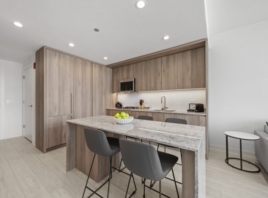Modern kitchen with light wood cabinets, built-in appliances, and a marble island with four dark gray chairs. A bowl of green apples sits on the island, and the kitchen is brightly lit with recessed lighting.