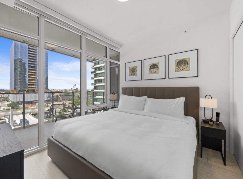 A modern bedroom with a large bed, white bedding, bedside tables with lamps, and three framed artworks above the headboard. Floor-to-ceiling windows reveal a city view with tall buildings and a balcony.