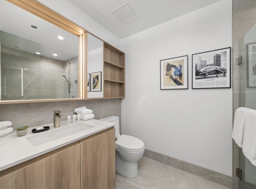 Modern bathroom with a light wood vanity, white countertop, and open shelving. There’s a toilet beneath two framed photos on the wall, light gray tile floors, and a glass-enclosed shower reflected in the large mirror.