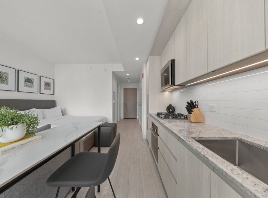 Modern studio apartment with a compact kitchen featuring light wood cabinets, a sink, stove, and microwave. There’s a dining area with two stools, a bed in the background, and minimalist decor on the walls.