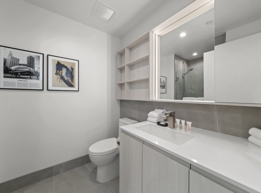 Modern bathroom with light-colored cabinets, a countertop with toiletries and towels, a toilet, wall art, open shelves, and a glass-enclosed shower reflected in a large mirror. The room is clean and minimalistic.
