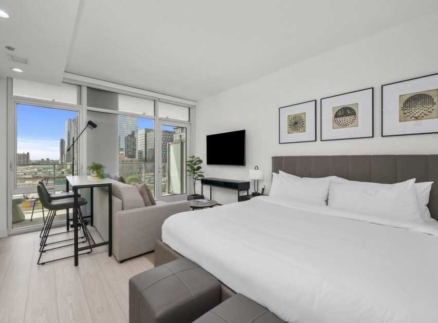 Modern studio apartment with a large bed, sofa, wall-mounted TV, desk, and dining table by floor-to-ceiling windows overlooking a city skyline. The decor is minimalist with framed art above the bed.