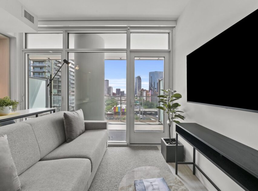 Modern living room with a gray sofa, large windows, and a cityscape view. A flat-screen TV is mounted on the wall across from a console table with a potted plant. Natural light fills the bright, minimalist space.