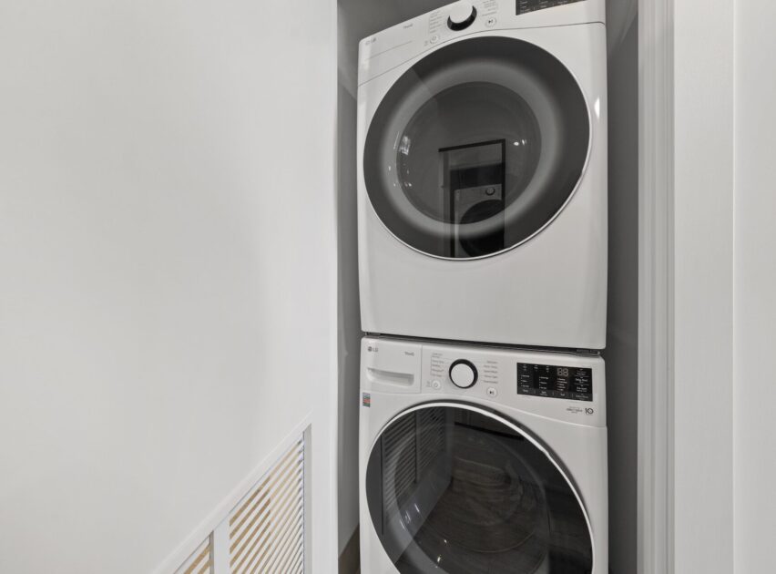 A stacked white washer and dryer set is placed in a narrow closet with white walls. The machines have round glass doors and digital controls. A vented panel is visible on the left side of the image.