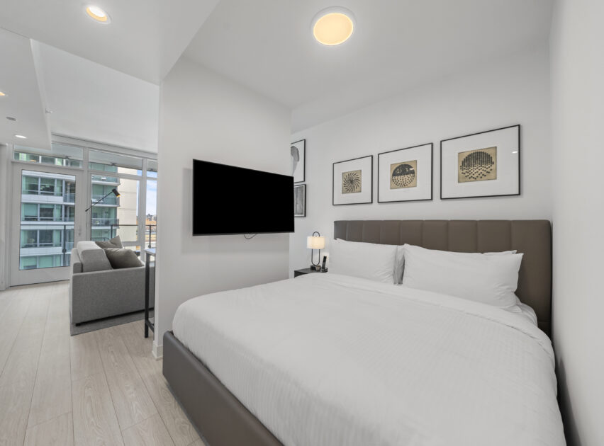 Modern bedroom with a neatly made bed, wall-mounted TV, three framed pictures above the headboard, a bedside lamp, and a view into a bright living area with large windows and a gray sofa.